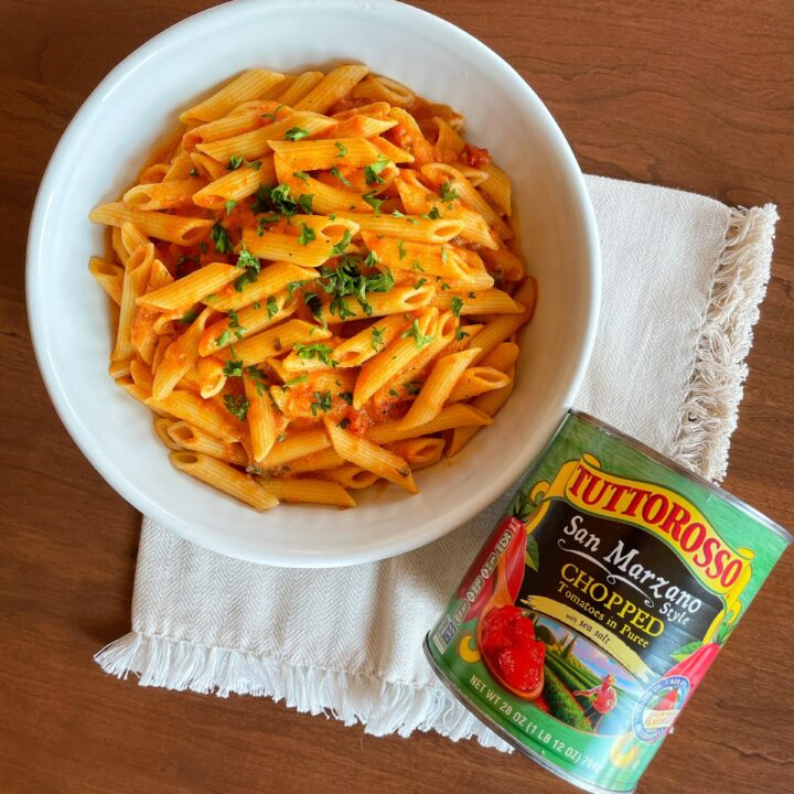 Bowl of penne pasta with dairy-free vodka sauce on a wood table with cream colored cloth and a can of Tuttorosso tomatoes next to it.